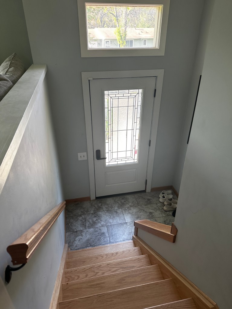 Interior view looking down the split-level stairwell at the new glass-insert front door, still in its original white before painting, showing how much more natural light reaches the landing