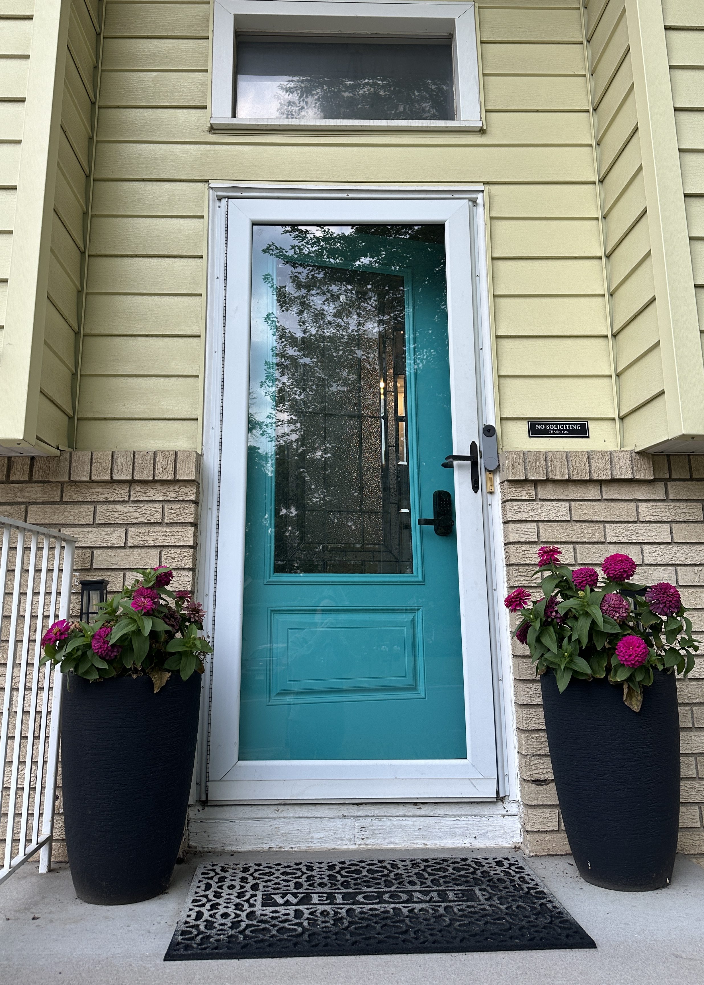 Straight-on exterior shot of the finished teal front door, flanked by potted flowers, with the house number and welcome mat in view